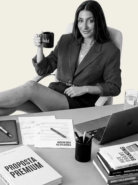 Brenda Scalco à mesa de trabalho, com uma caneca na mão e os papéis da proposta premium sobre a mesa